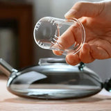 Hand holding a measuring cup next to an elegant glass teapot with a modern black handle.
